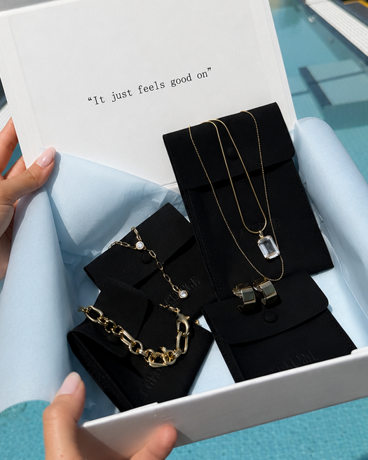 A white jewelry gift box held by two hands over a rooftop pool, containing gold-toned necklaces and bracelets displayed on black velvet stands. The box lid has the text 'It just feels good us' printed on it, with a city skyline and pool in the background.