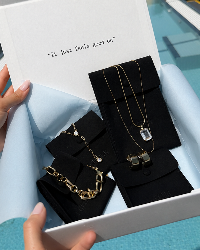A white jewelry gift box held by two hands over a rooftop pool, containing gold-toned necklaces and bracelets displayed on black velvet stands. The box lid has the text 'It just feels good us' printed on it, with a city skyline and pool in the background.