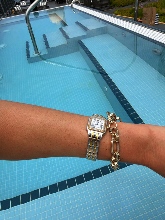 A gold-tone watch with a square face and link bracelet worn on a person's wrist, alongside a chunky gold chain bracelet, with a blue tiled swimming pool in the background.